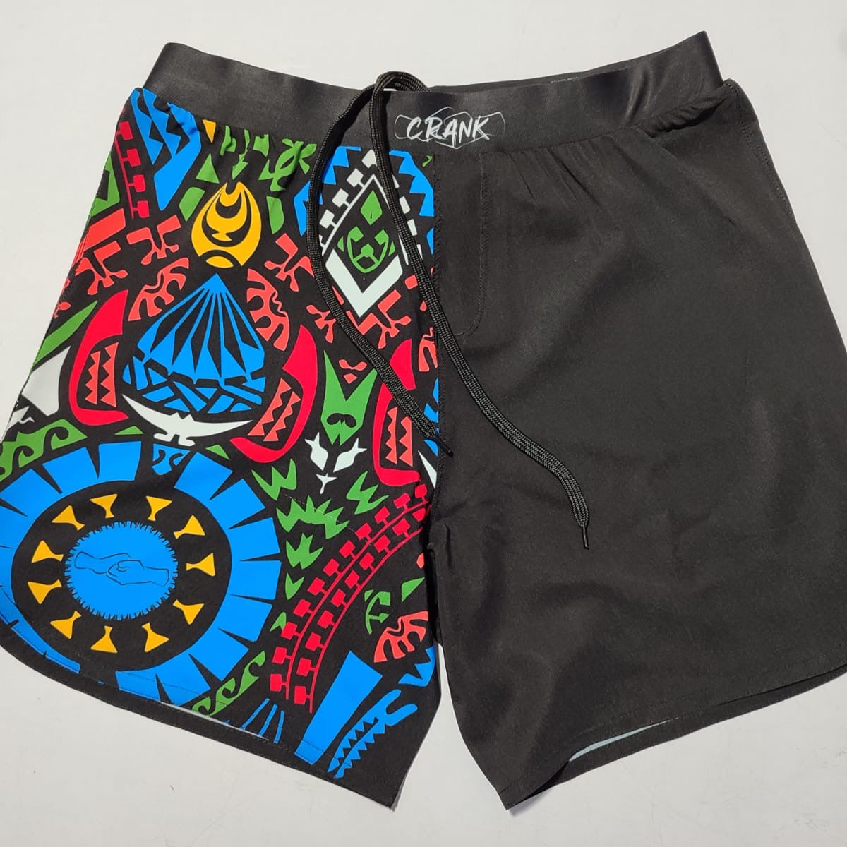 A pair of grappling shorts with a colorful Aztec pattern on the left side and solid black on the right side, with the brand 'CRANK' visible on the waistband.